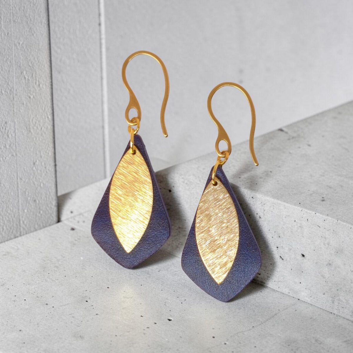 Boucles d'oreilles forme larme en cuir sauvage bleu, taille réduite, finition aniline et pendants dorés brossés, vues de face, suspendues à des boucles dorées sur béton