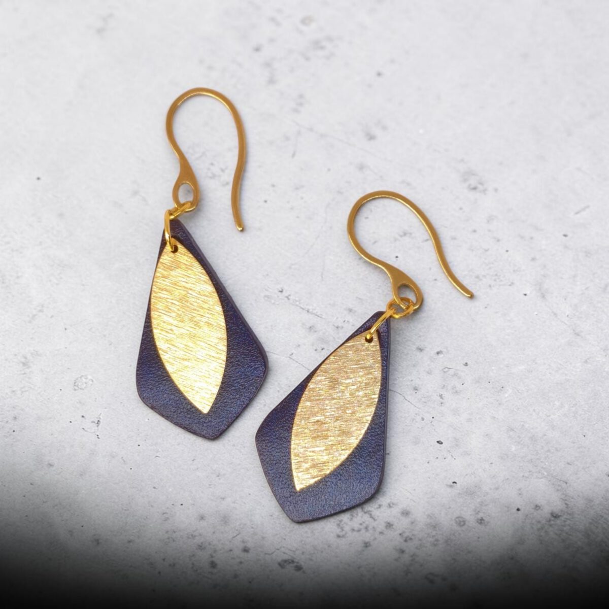 Boucles d'oreilles forme larme en cuir sauvage bleu, taille réduite, finition aniline et pendants dorés brossés, vues de face, suspendues à des boucles dorées, fond béton dégradé
