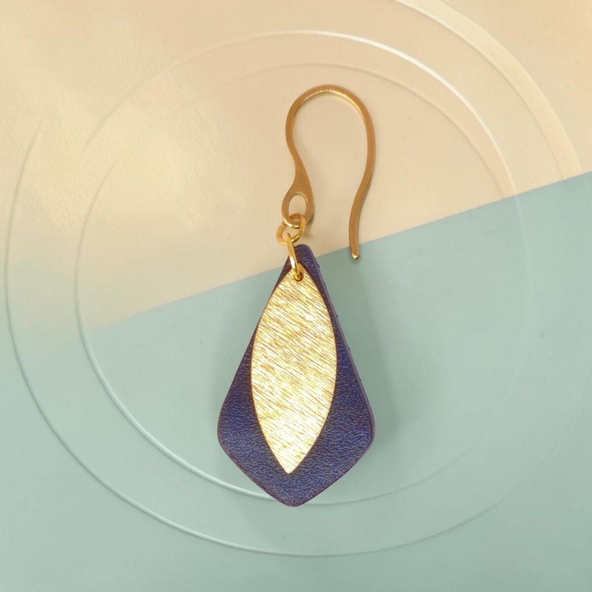 Boucle d'oreille forme larme en cuir sauvage bleu, taille réduite, finition aniline et pendant doré brossé, vue de face, suspendue à une boucle dorée, fond bicolore