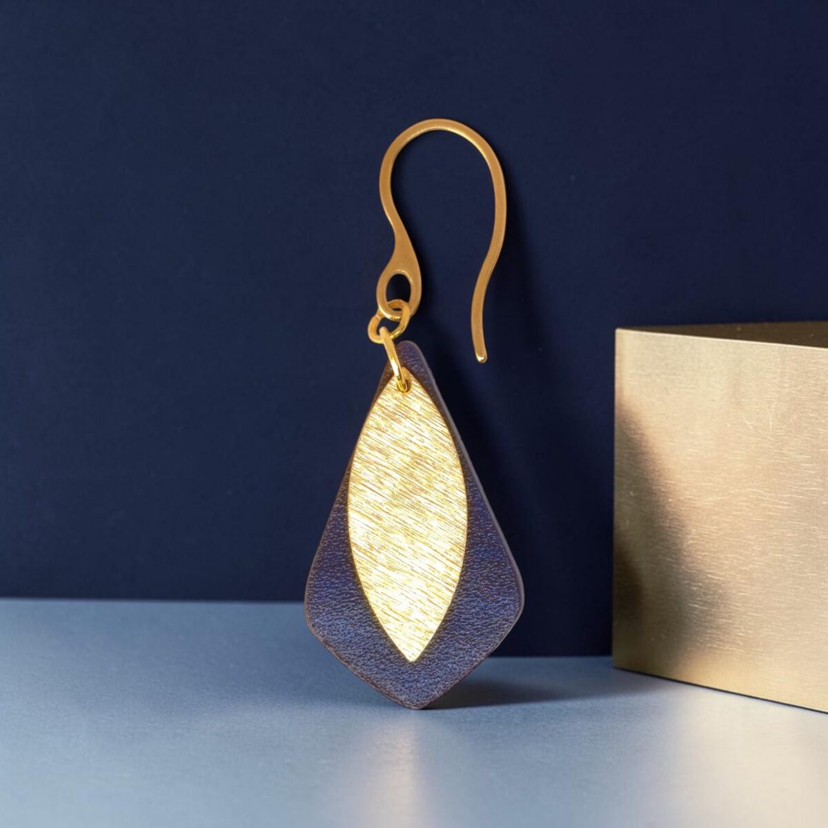 Boucle d'oreille forme larme en cuir sauvage bleu, taille réduite, finition aniline et pendant doré brossé, vue de face, suspendue à une boucle dorée, fond design