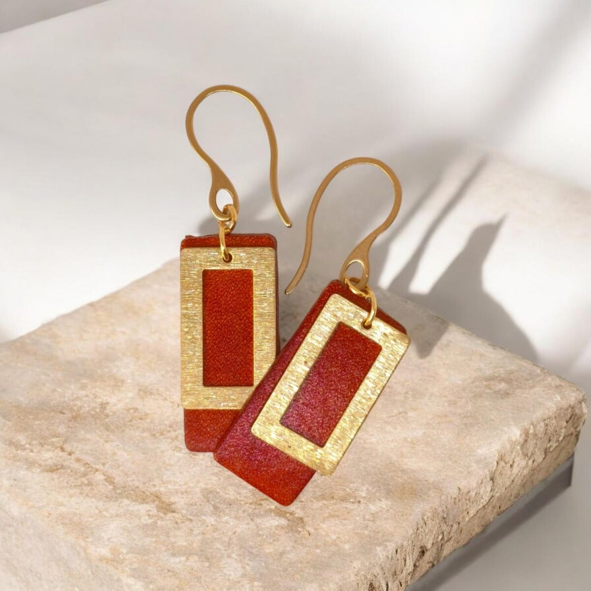 Boucles d'oreilles rectangulaires en cuir teinté cognac, finition lisse et pendant brossé doré, vues de face se chevauchant, suspendues à des boucles dorées, sur pierre