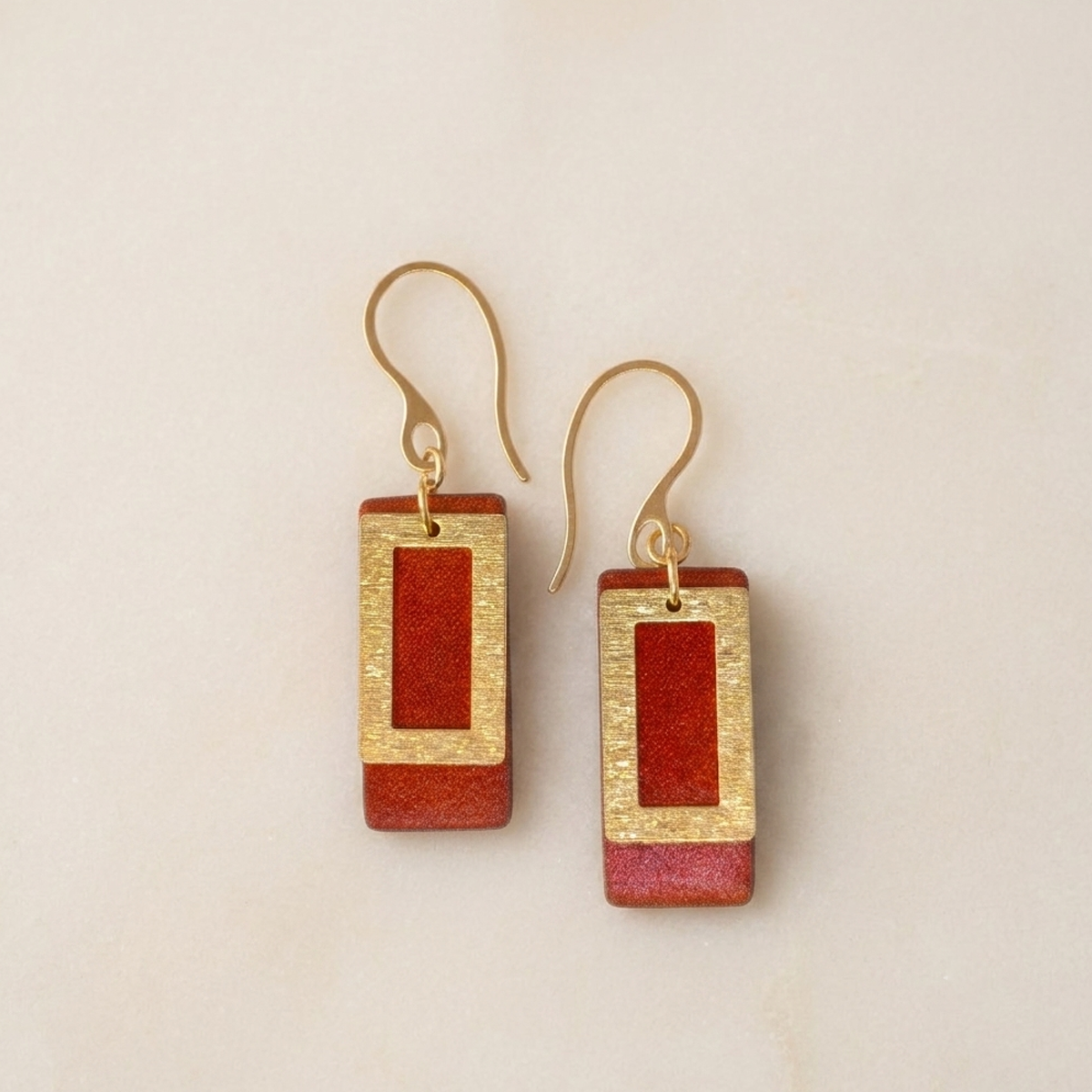 Boucles d'oreilles rectangulaires en cuir teinté cognac, finition lisse et pendant brossé doré, vues de face, suspendues à des boucles dorées, fond sable