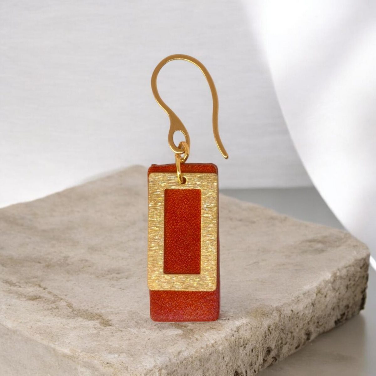 Boucle d'oreille rectangulaire en cuir teinté cognac, finition lisse et pendant brossé doré, vue de face, suspendue à une boucle dorée, sur pierre