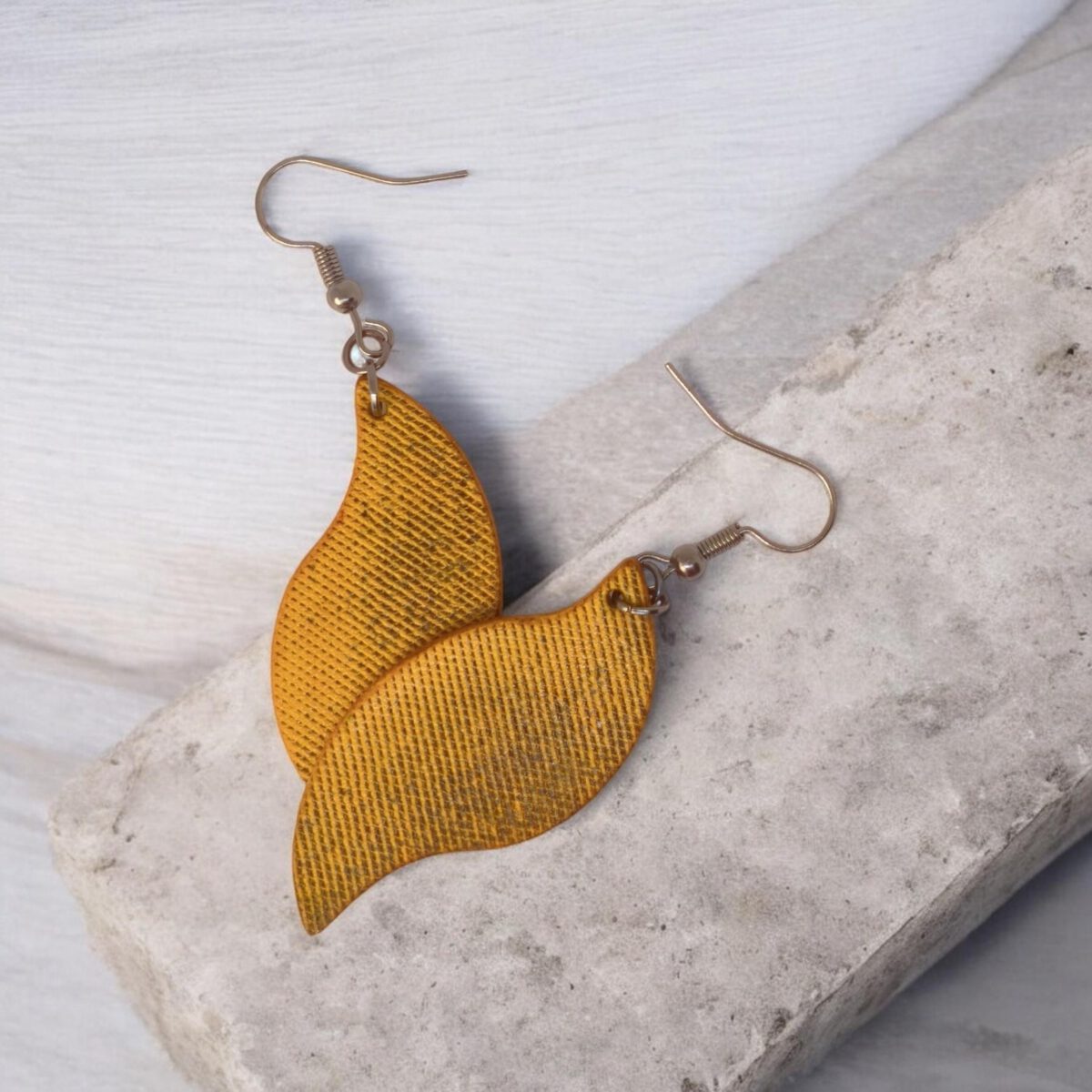 Boucles d'oreilles forme feuille asymétrique en cuir teinté jaune, texturage lime fine vieillie, vues de face se chevauchant, suspendues à des boucles gun métal, sur pierre