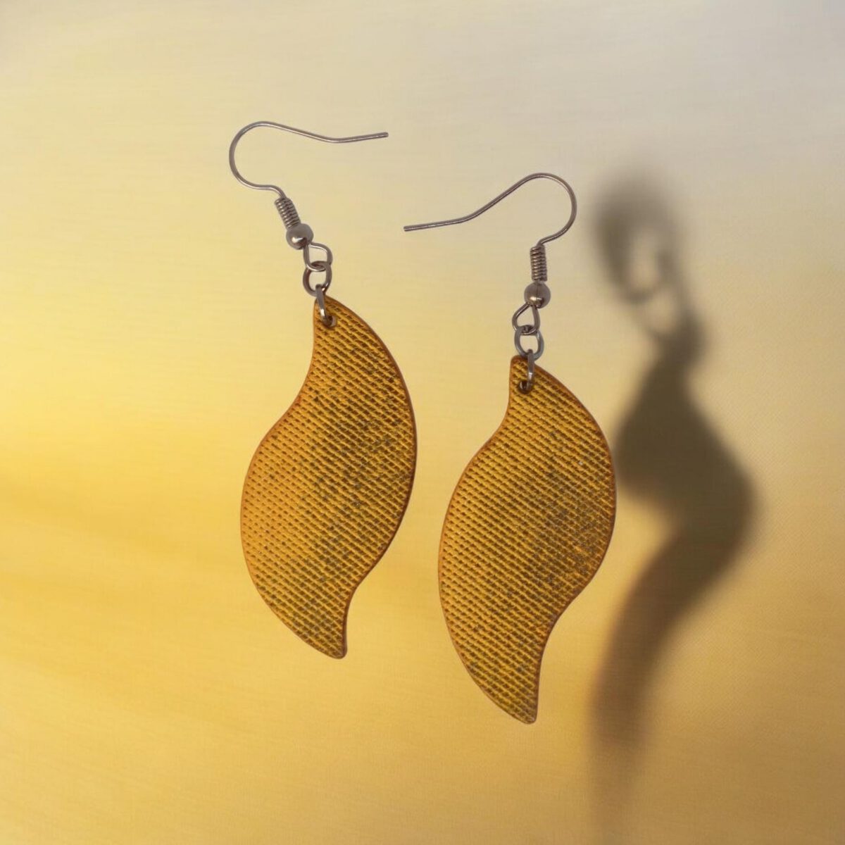 Boucles d'oreilles forme feuille asymétrique en cuir teinté jaune, texturage lime fine vieillie, vues de face, suspendues à des boucles gun métal, fond jaune