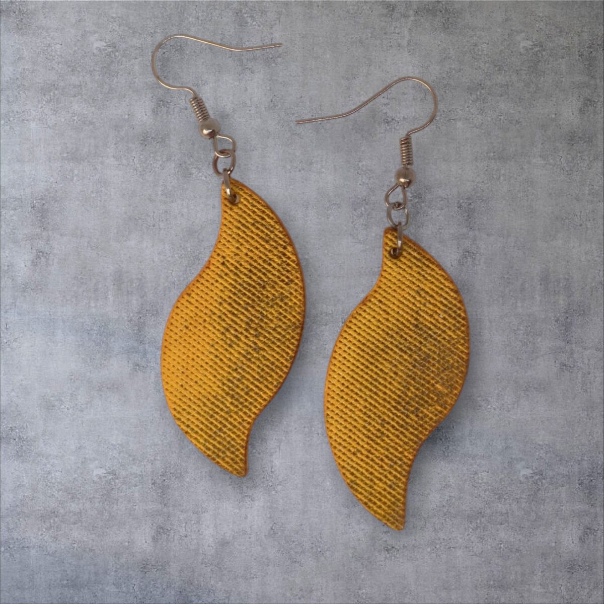 Boucles d'oreilles forme feuille asymétrique en cuir teinté jaune, texturage lime fine vieillie, vues de face, suspendues à des boucles gun métal, fond gris