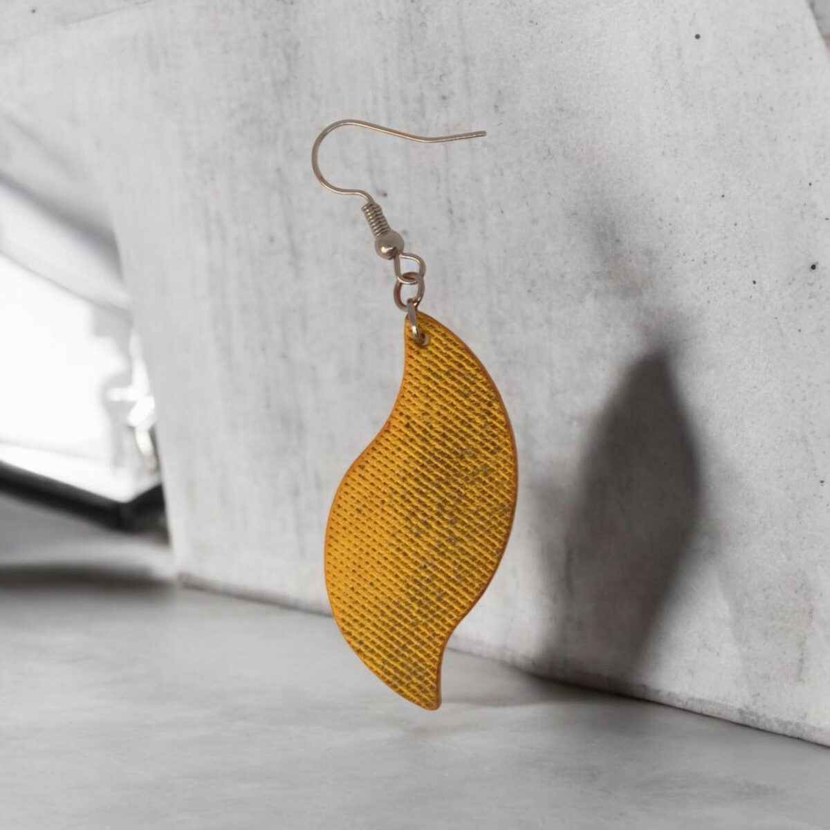 Boucle d'oreille forme feuille asymétrique en cuir teinté jaune, texturage lime fine vieillie, vue de face, suspendue à une boucle gun métal, fond béton