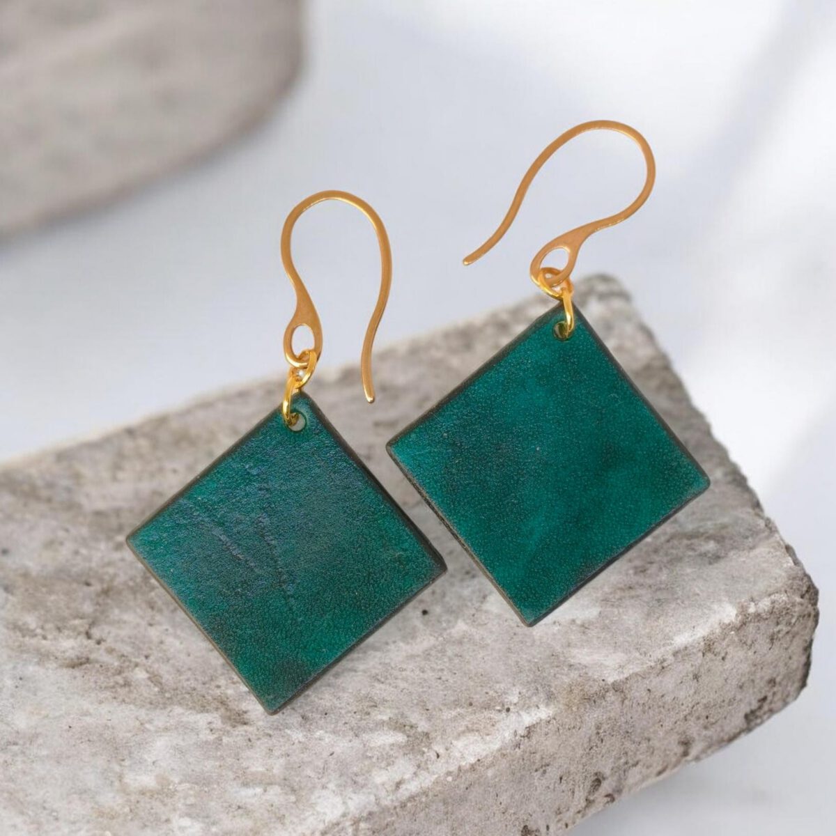 Boucles d'oreilles carrées en cuir teinté vert d'eau, finition lisse, vues de face, suspendues à des boucles dorées, sur pierre