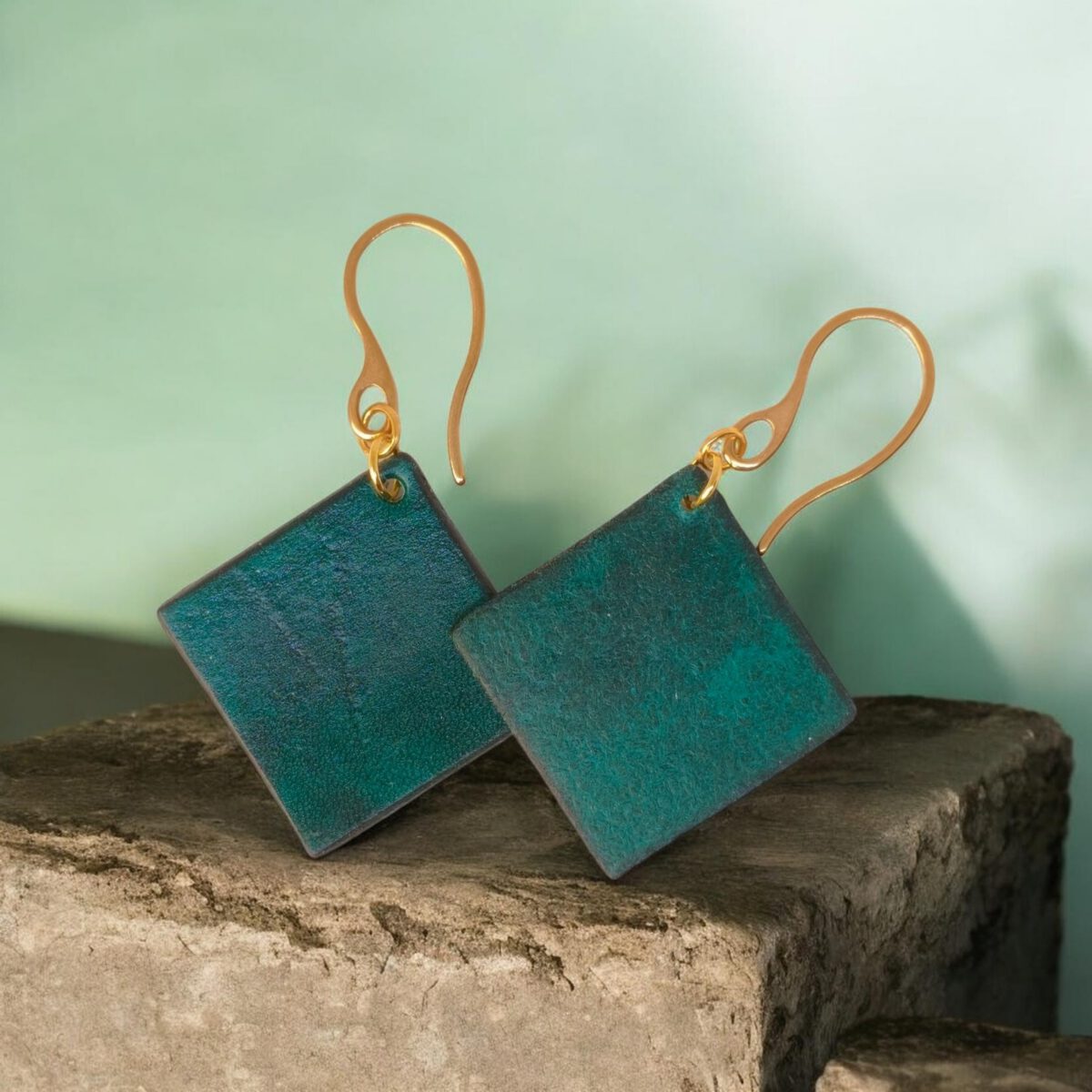 Boucles d'oreilles carrées en cuir teinté vert d'eau, finition lisse, vues de face se chevauchant, suspendues à des boucles dorées, sur pierre