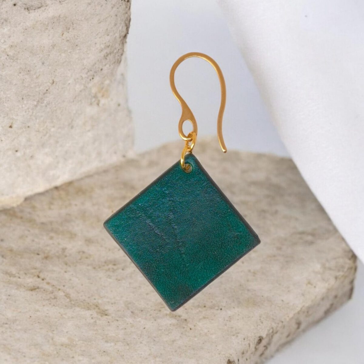 Boucle d'oreille carrée en cuir teinté vert d'eau, finition lisse, vue de face, suspendue à une boucle dorée, sur pierre