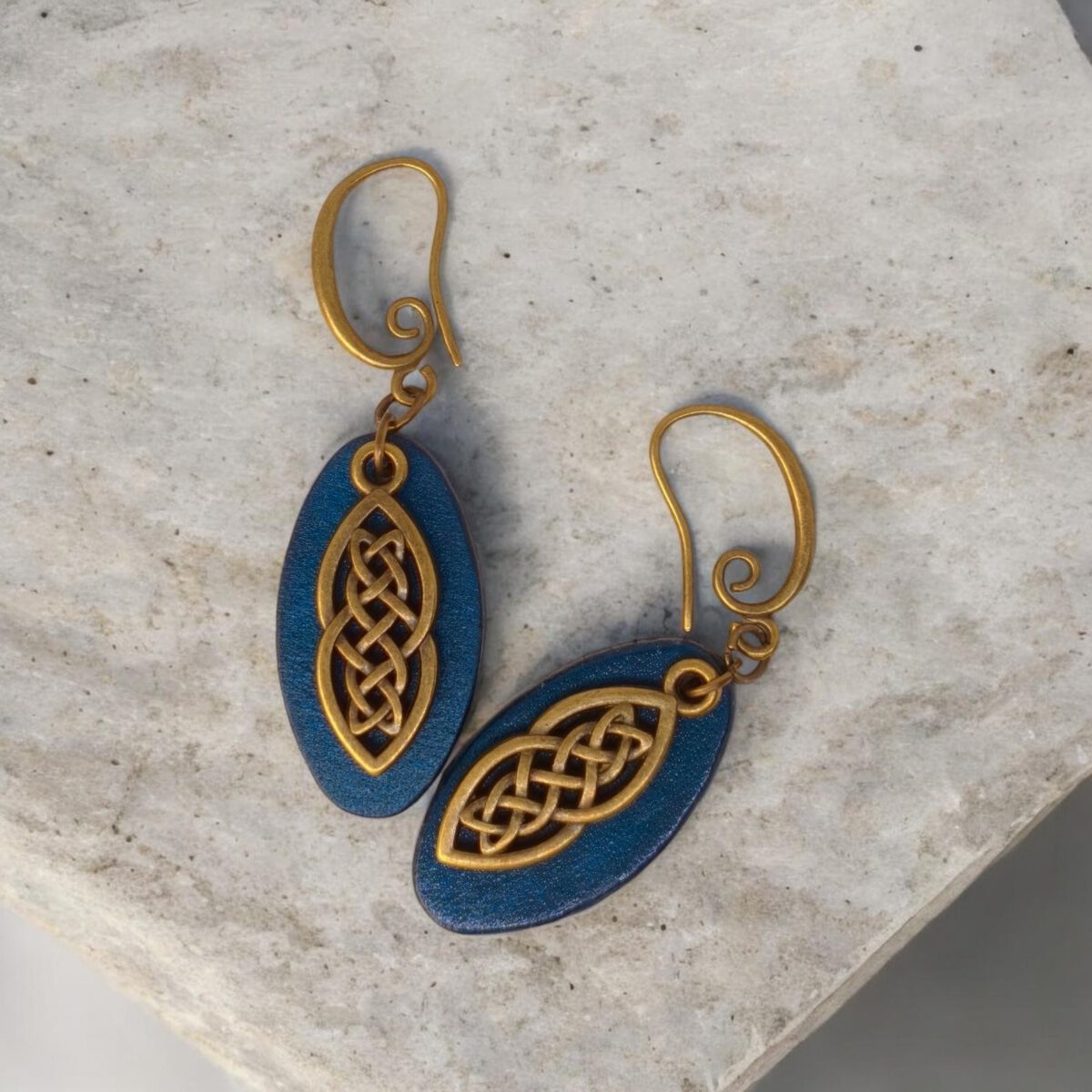 Boucles d'oreilles ovales en cuir teinté bleu, finition lisse et pendant celtique, vues de face, suspendues à des boucles couleur bronze sur pierre