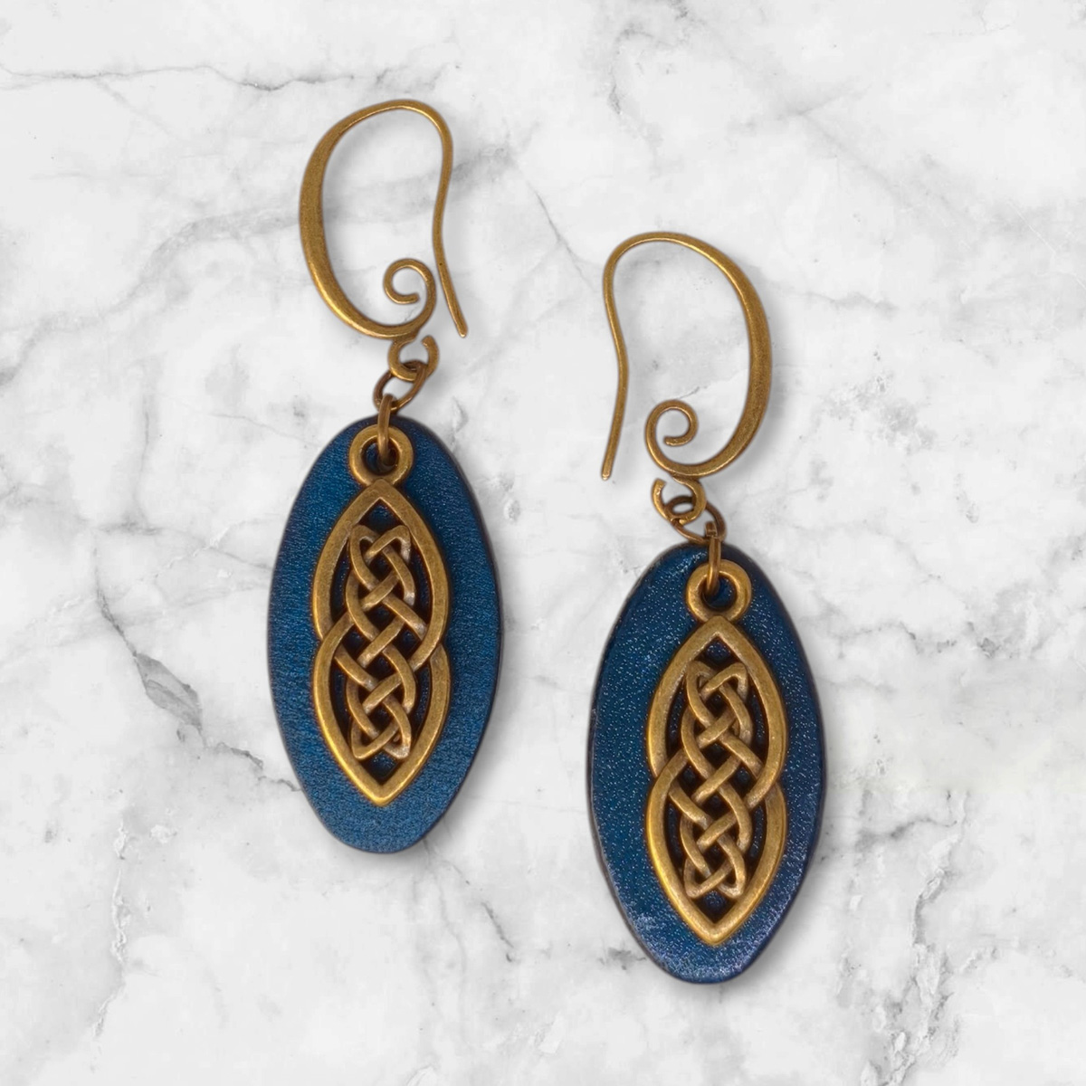 Boucles d'oreilles ovales en cuir teinté bleu, finition lisse et pendant celtique, vues de face, suspendues à des boucles couleur bronze sur marbre