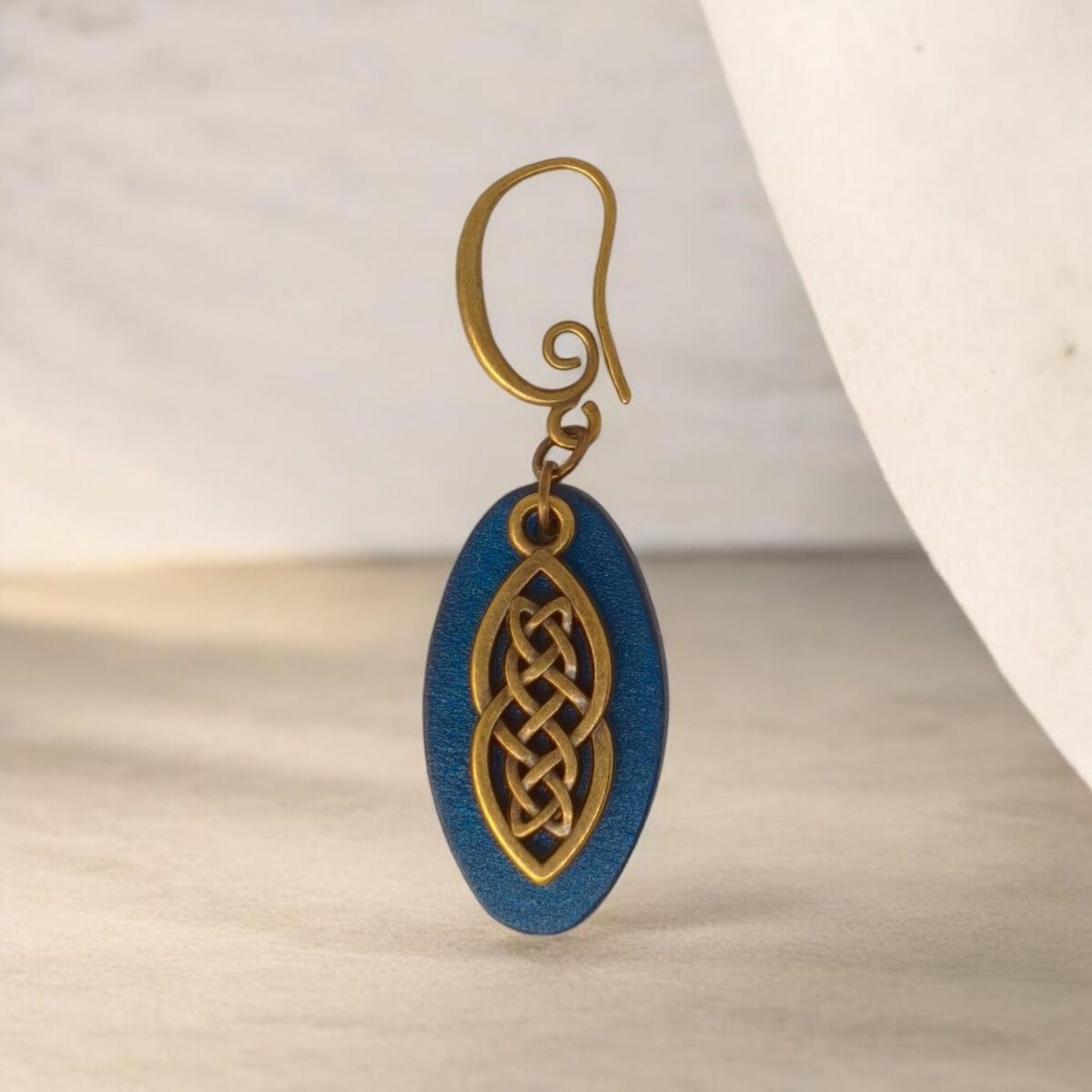 Boucle d'oreille ovale en cuir teinté bleu, finition lisse et pendant celtique, vue de face, suspendue à une boucle couleur bronze, fond design