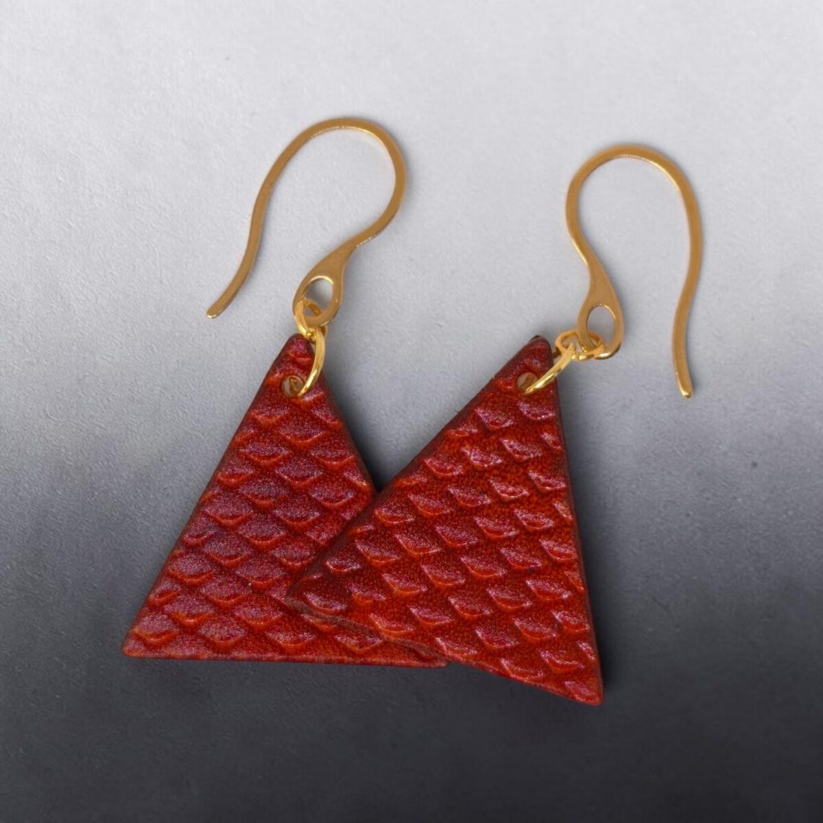Boucles d'oreilles forme triangulaire en cuir teinture cognac, texturage poisson fin, vues de face se chevauchant, suspendues à des boucles dorées fond noir