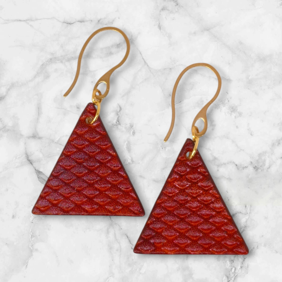 Boucles d'oreilles forme triangulaire en cuir teinture cognac, texturage poisson fin, vues de face, suspendues à des boucles dorées sur marbre