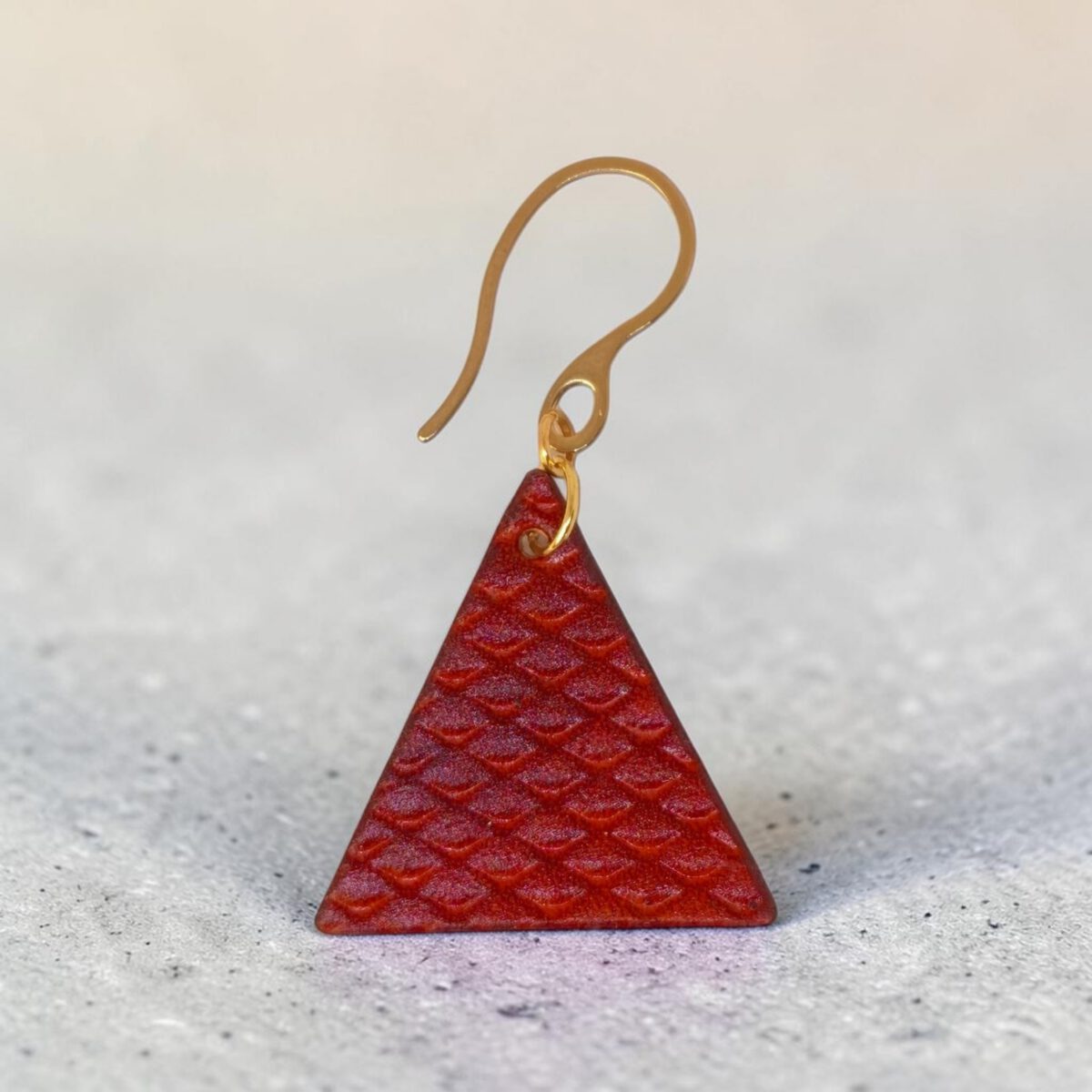 Boucle d'oreille forme triangulaire en cuir teinture cognac, texturage poisson fin, vue de face, suspendue à une boucle dorée, sur béton