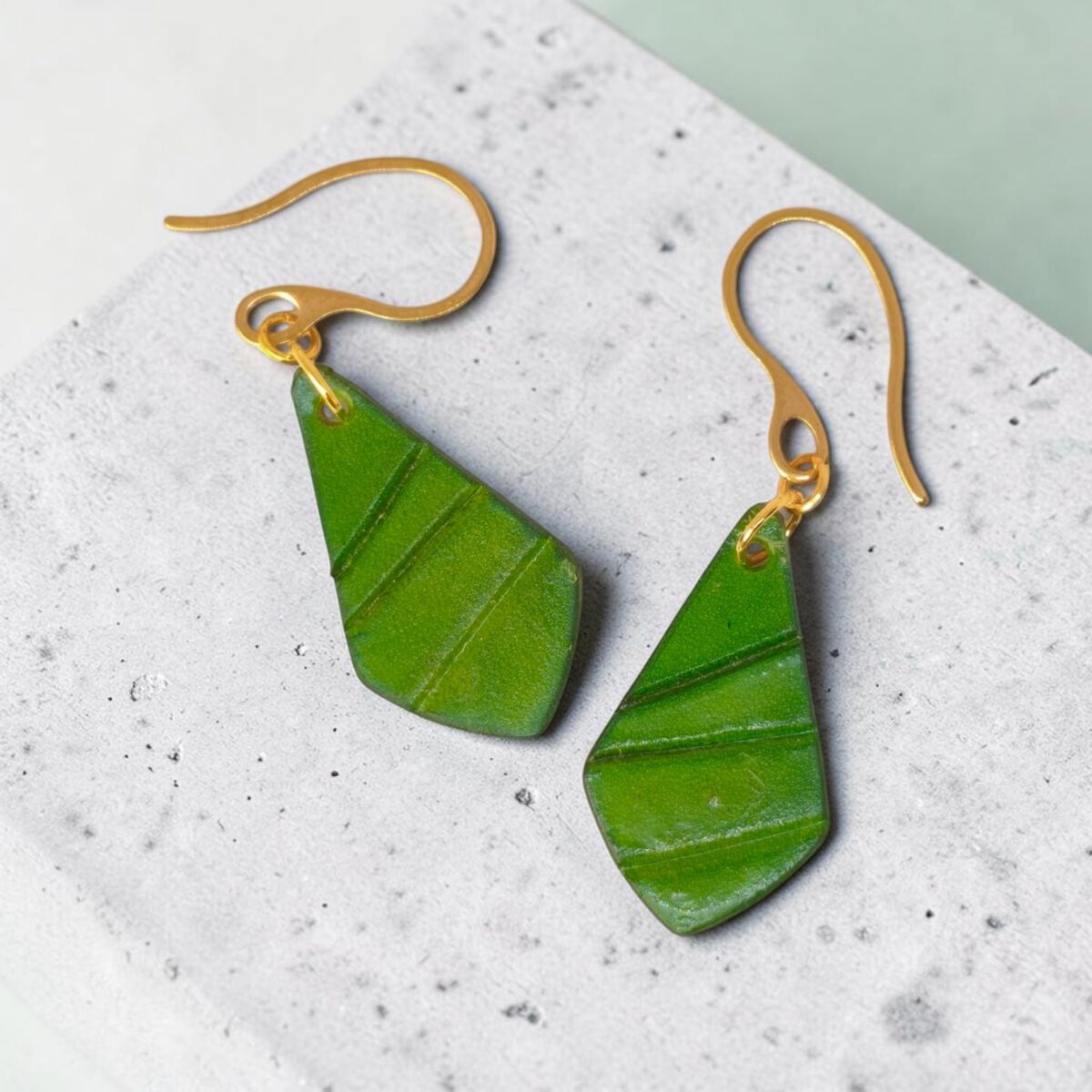 Boucles d'oreilles forme larme en cuir teinture vert forêt, taille réduite, finition galet, vues de face, suspendues à des boucles dorées sur béton