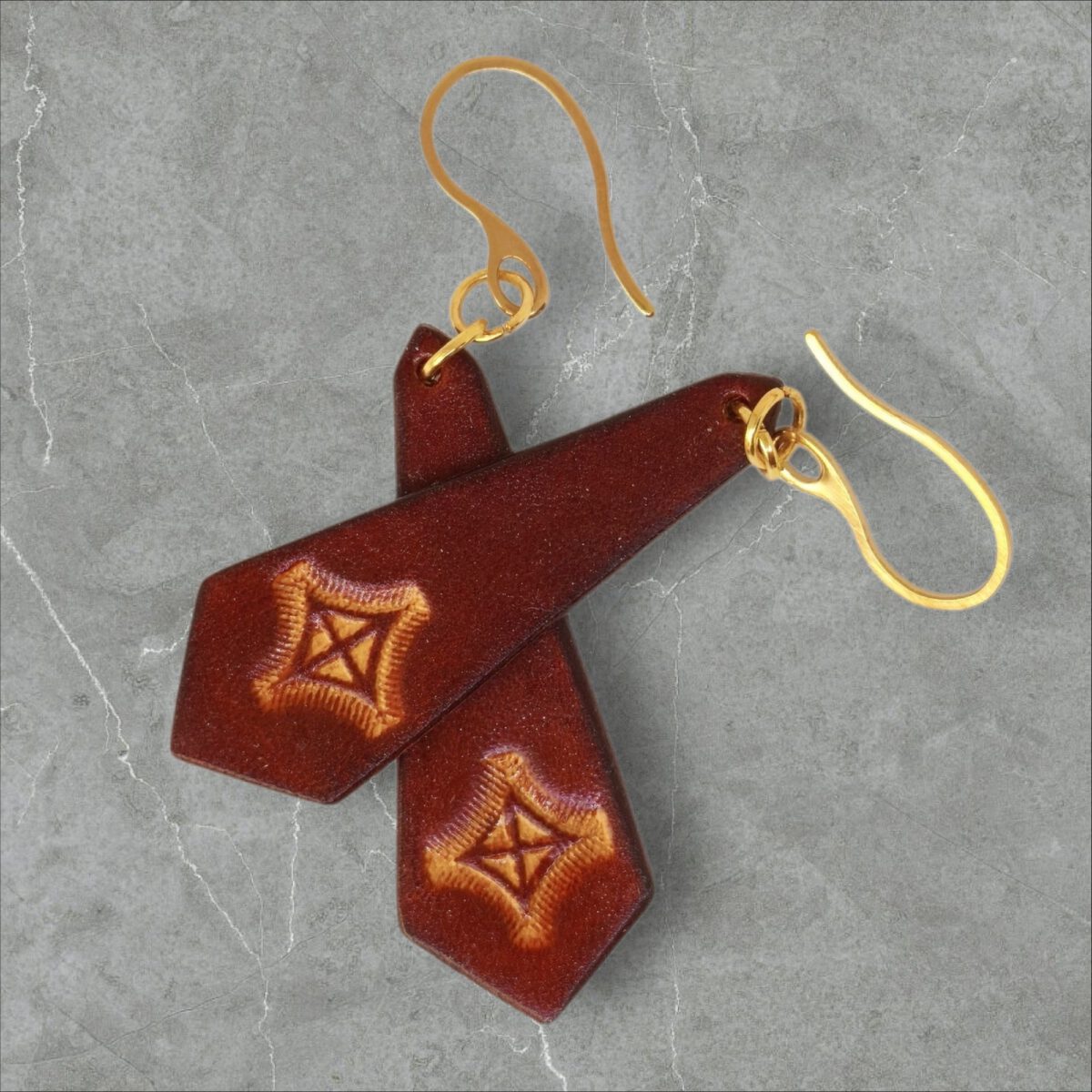 Boucles d'oreilles cravates en cuir teinte cognac, estampage croix teintée jaune, vues de face se chevauchant, suspendues à des boucles dorées sur fond gris