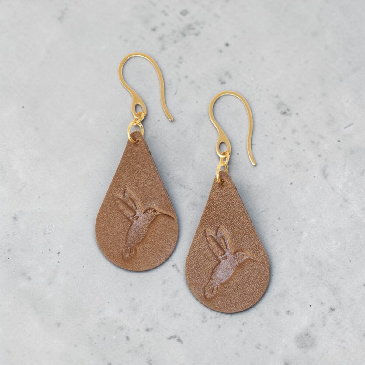 Boucles d'oreilles forme goutte d'eau en cuir marron, estampages colibri, vues de face, suspendues à des boucles dorées sur béton