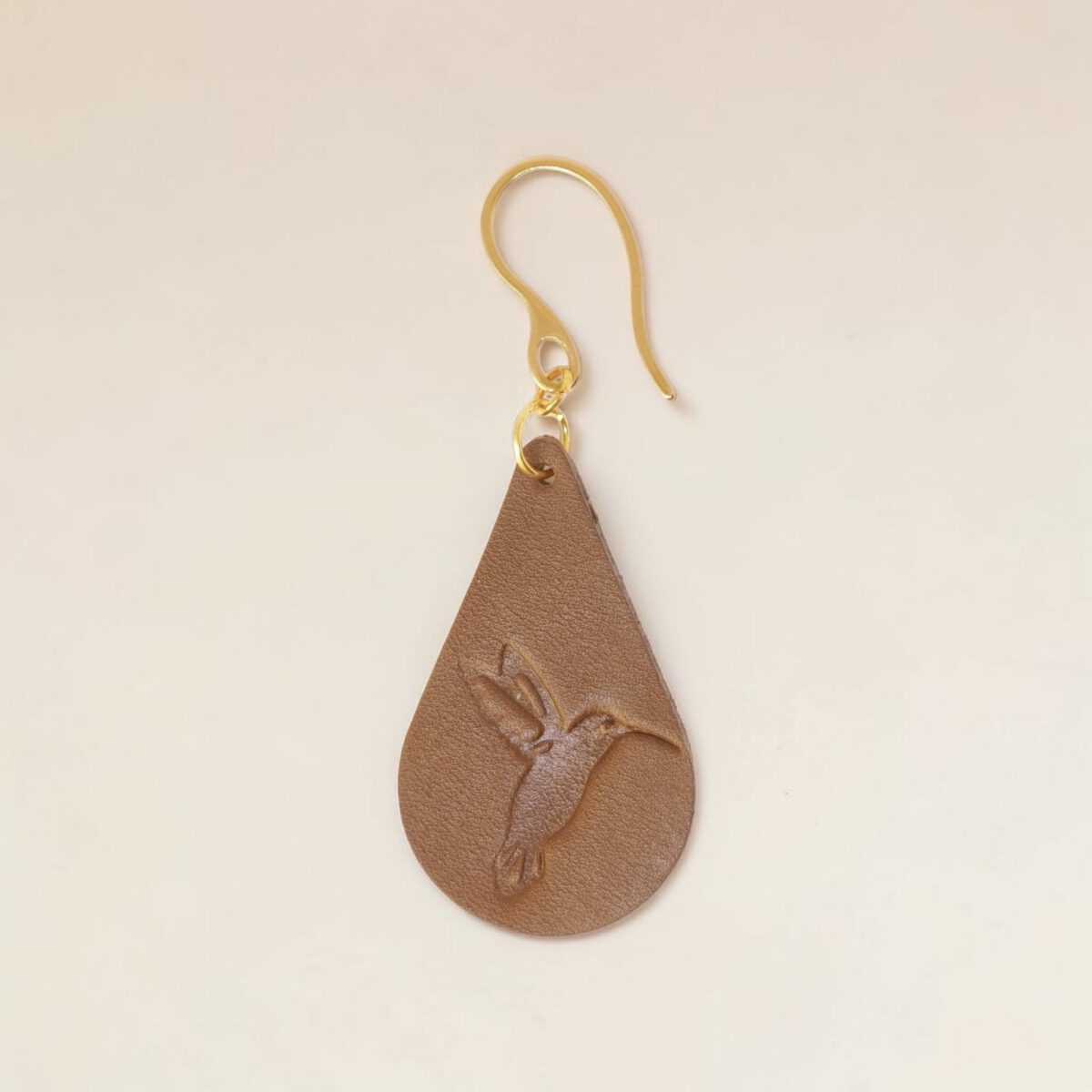Boucle d'oreille forme goutte d'eau en cuir marron, estampage colibri, vue de face, suspendue à une boucle dorée, fond beige