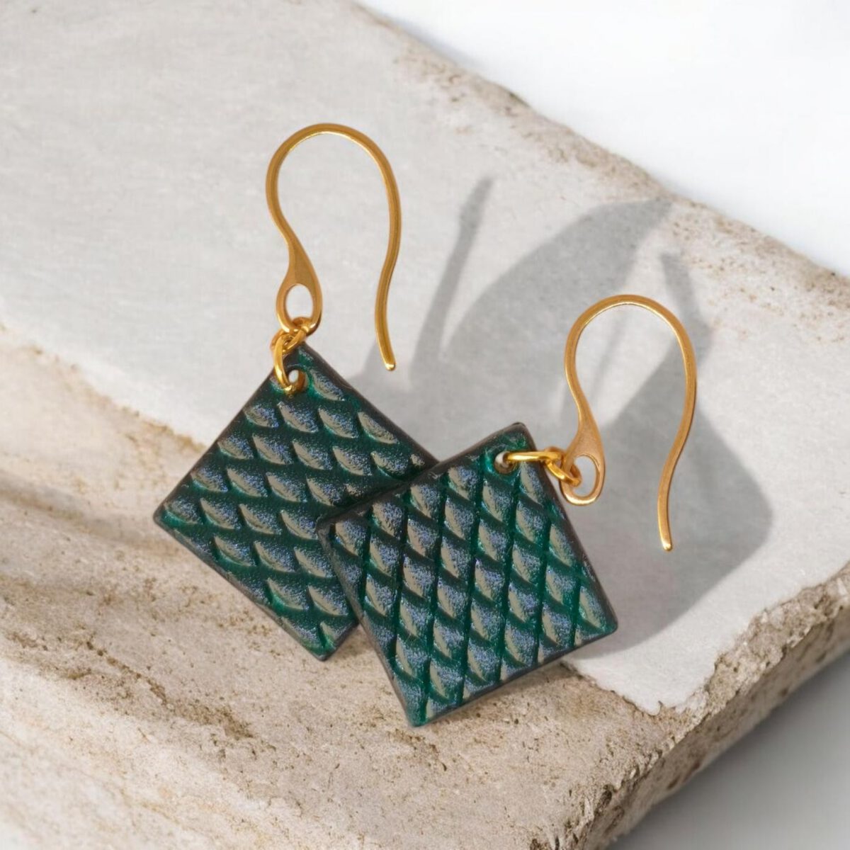 Boucles d'oreilles carrées en cuir teinté vert d'eau, texturage poisson, vues de face se chevauchant, suspendues à des boucles dorées, sur pierre