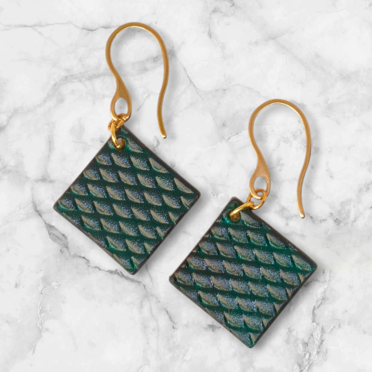 Boucles d'oreilles carrées en cuir teinté vert d'eau, texturage poisson, vues de face, suspendues à des boucles dorées, fond marbre