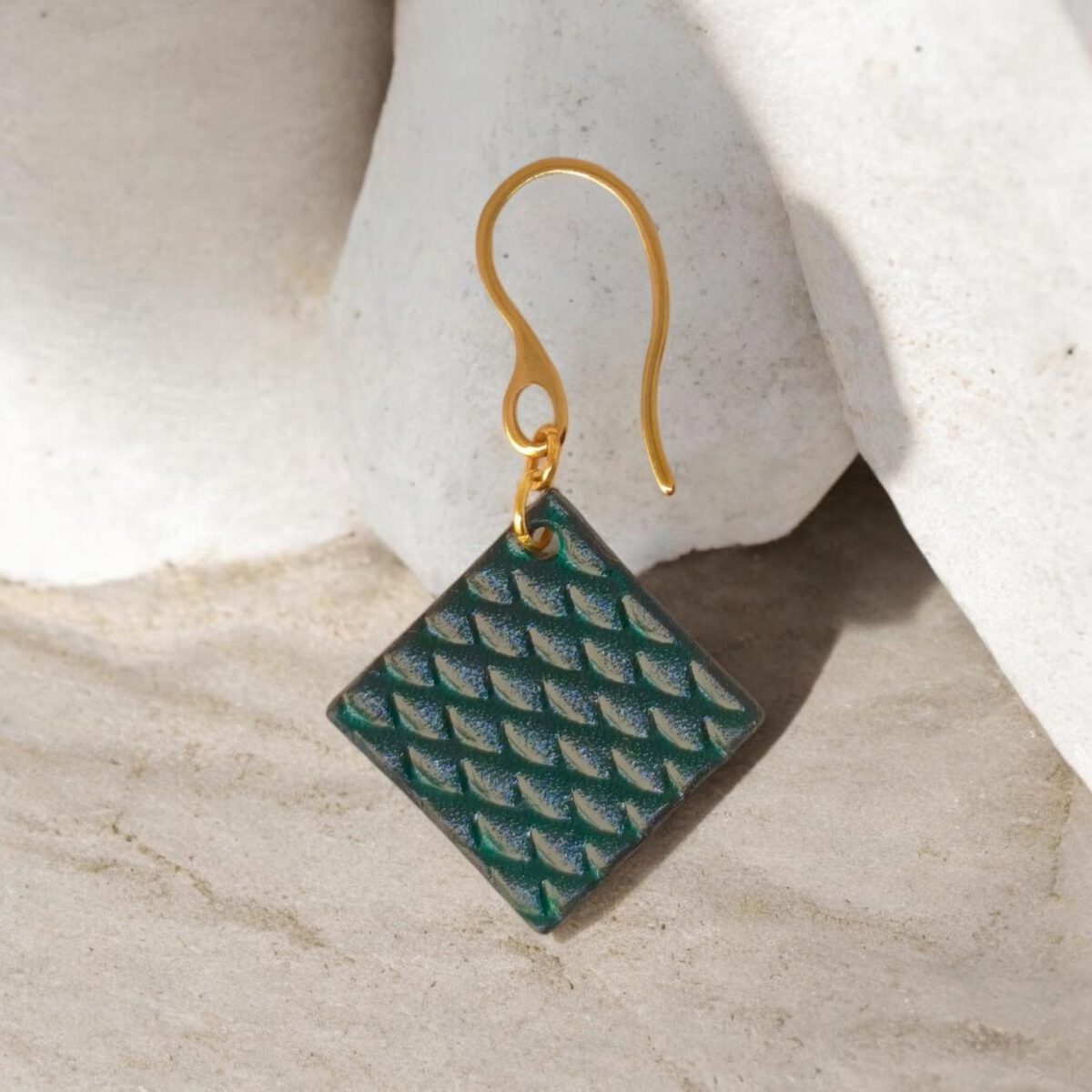 Boucle d'oreille carrée en cuir teinté vert d'eau, texturage poisson, vue de face, suspendue à une boucle dorée, sur pierre