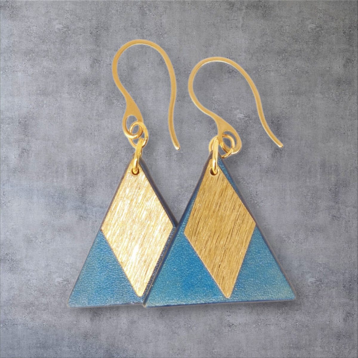 Boucles d'oreilles forme triangulaire en cuir teinture bleue, finition lisse et pendants losange dorés brossés vues de face se chevauchant, suspendues à des boucles dorées sur fond gris
