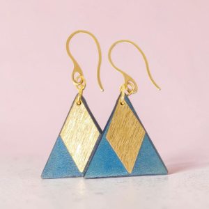 Boucles d'oreilles forme triangulaire en cuir teinture bleue, finition lisse et pendants losange dorés brossés vues de face se chevauchant, suspendues à des boucles dorées sur béton ciré