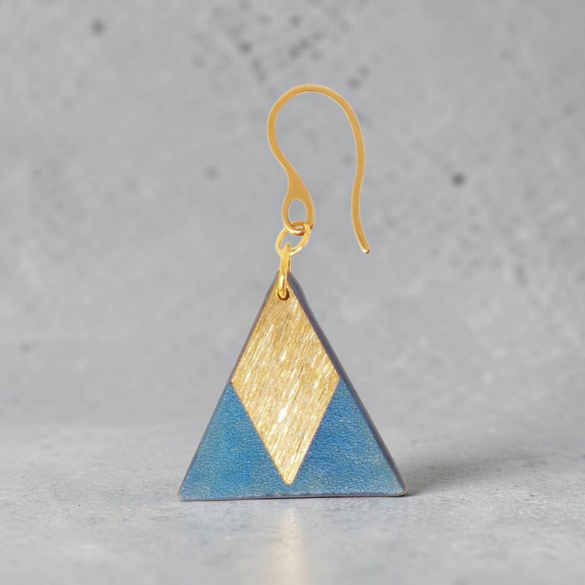 Boucle d'oreille forme triangulaire en cuir teinture bleue, finition lisse et pendant losange doré brossé, vue de face, suspendue à une boucle dorée, sur béton ciré