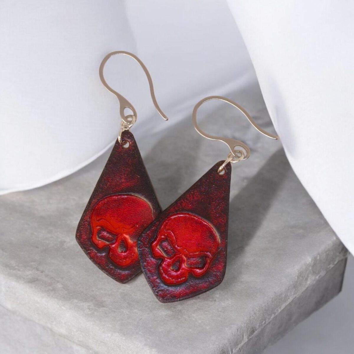Boucles d'oreilles forme larme en cuir teinture rouge et noire, estampage tête de mort, vues de face se chevauchant, suspendues à des boucles argentées, sur pierre