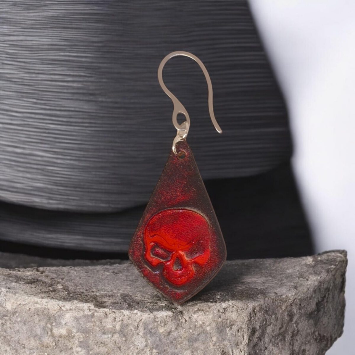 Boucle d'oreille forme larme en cuir teinture rouge et noire, estampage tête de mort, vue de face, suspendue à une boucle argentée, sur pierre