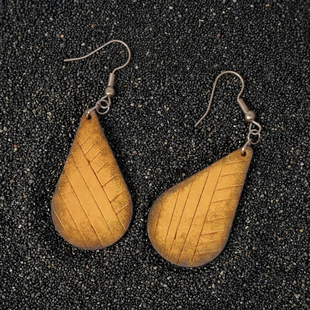 Boucles d'oreilles forme goutte d'eau en cuir teinture jaune dégradée, finition griffée, vues de face, suspendues à des boucles argentées sur sable noir