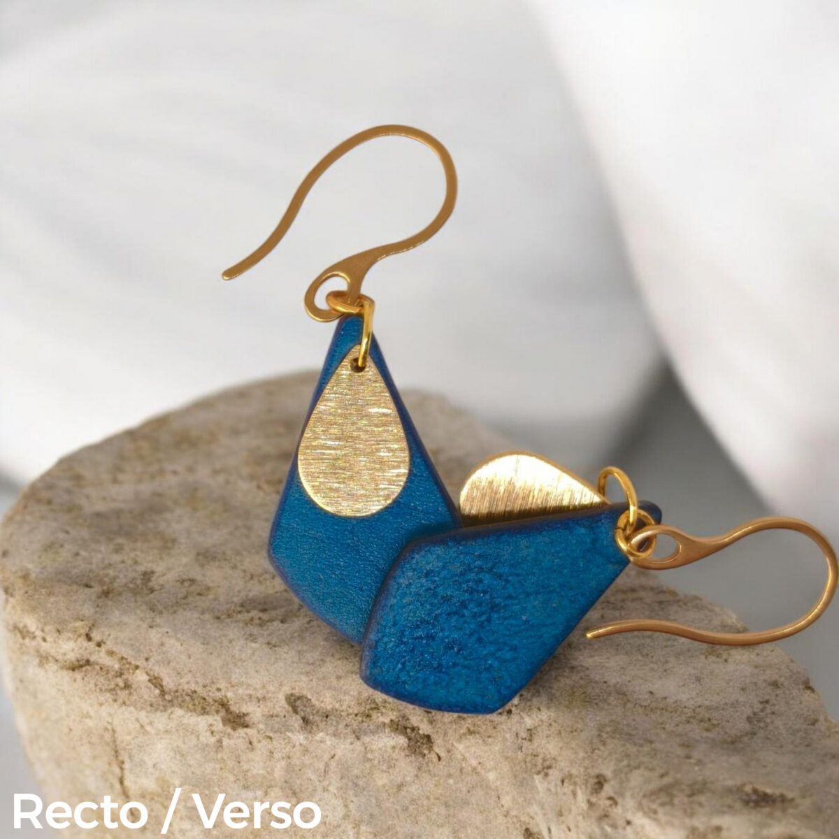 Boucles d'oreilles forme larme en cuir teinture bleue, finition lisse et pendants dorés brossés, vues recto - verso se chevauchant, suspendues à des boucles dorées sur pierre