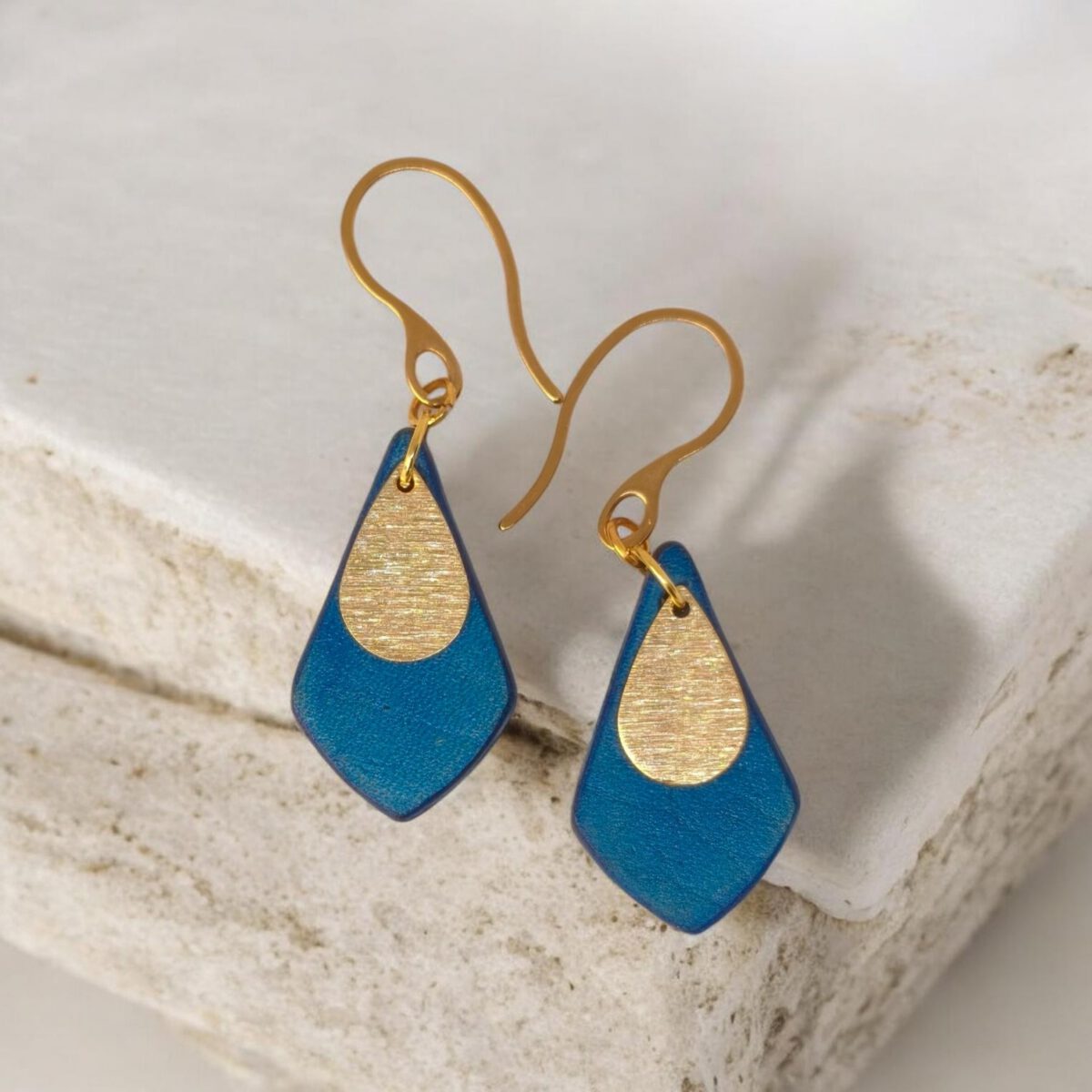 Boucles d'oreilles forme larme en cuir teinture bleue, finition lisse et pendants dorés brossés, vues de face, suspendues à des boucles dorées sur pierre