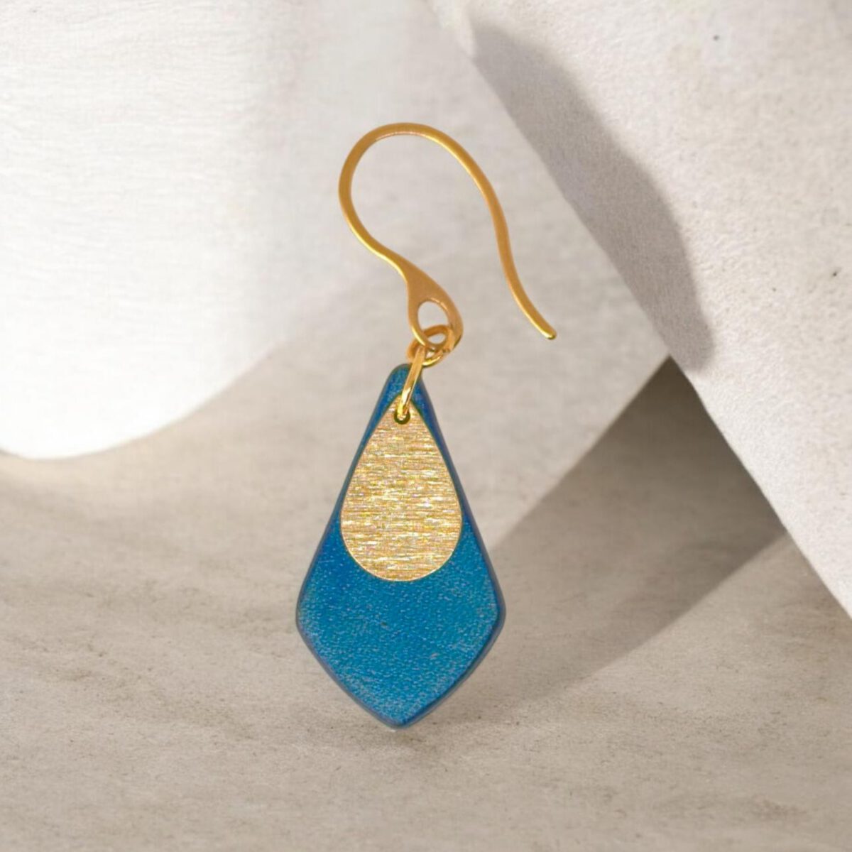 Boucle d'oreille forme larme en cuir teinture bleue, finition lisse et pendant doré brossé, vue de face, suspendue à une boucle dorée, fond sablé