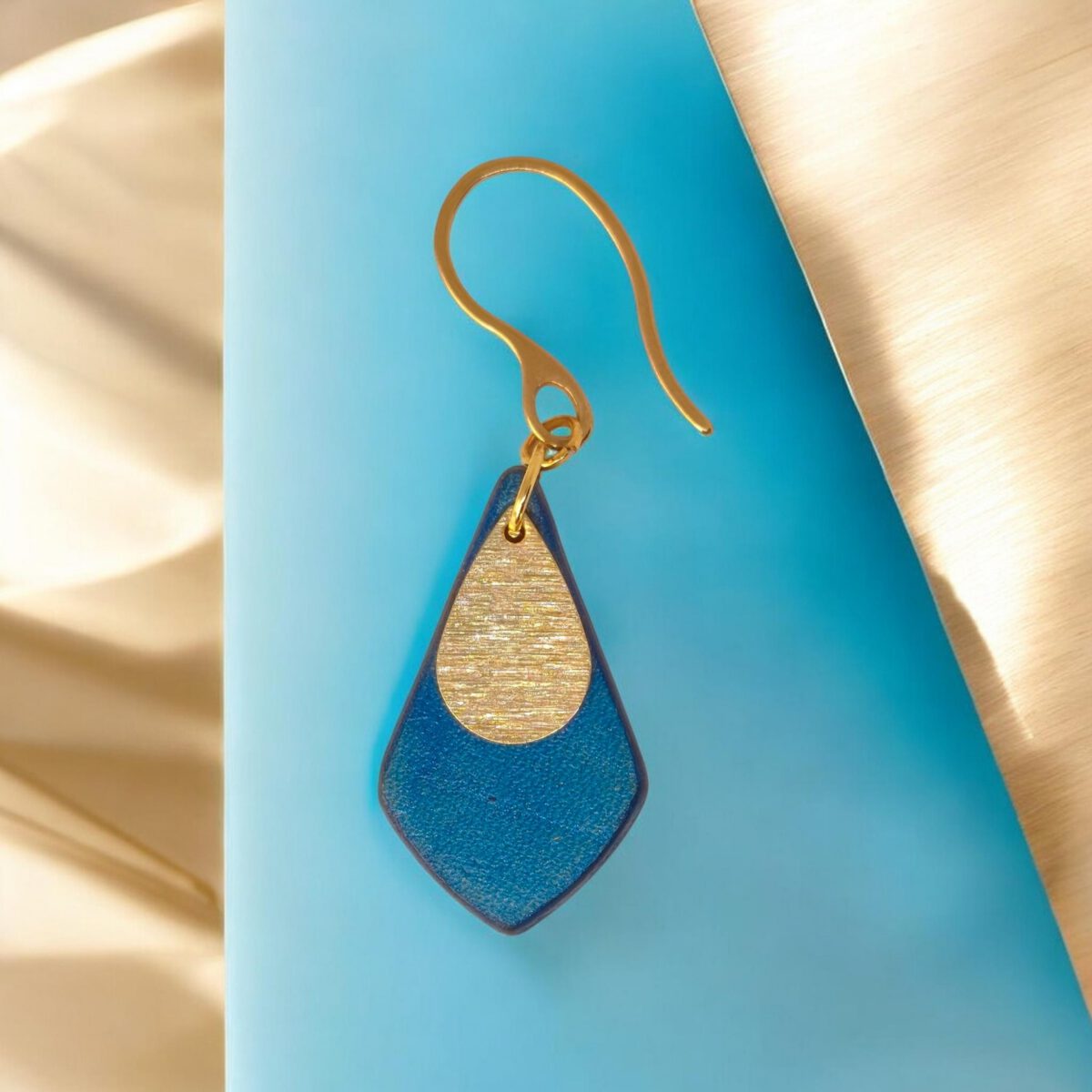 Boucle d'oreille forme larme en cuir teinture bleue, finition lisse et pendant doré brossé, vue de face, suspendue à une boucle dorée, fond design