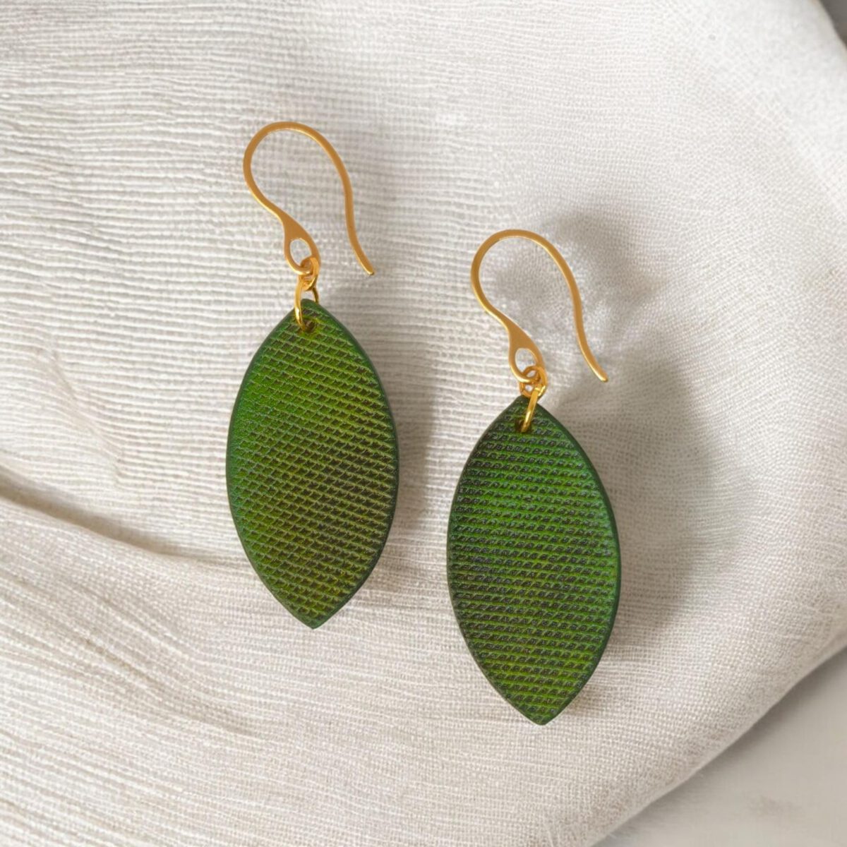 Boucles d'oreilles forme poisson en cuir teinture vert forêt, texturage lime épaisse, vues de face, suspendues à des boucles dorées sur lin