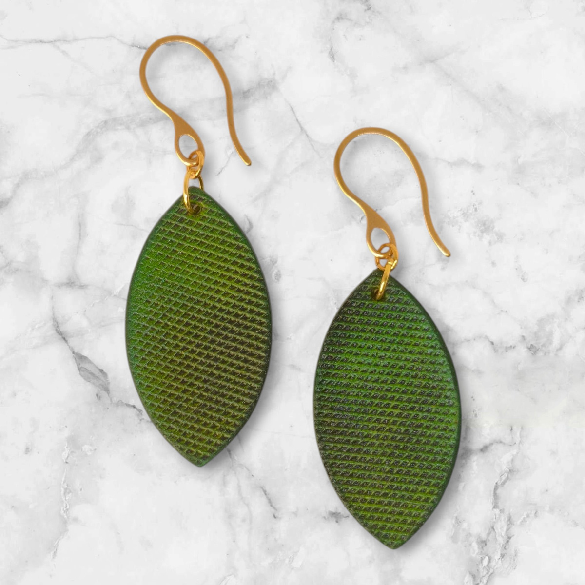 Boucles d'oreilles forme poisson en cuir teinture vert forêt, texturage lime épaisse, vues de face, suspendues à des boucles dorées sur marbre