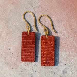 Boucles d'oreilles rectangulaires en cuir teinté cognac, texturage quadrillé, vues de face, suspendues à des boucles dorées, sur béton