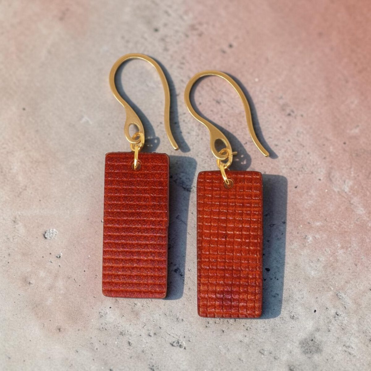 Boucles d'oreilles rectangulaires en cuir teinté cognac, texturage quadrillé, vues de face, suspendues à des boucles dorées, sur béton