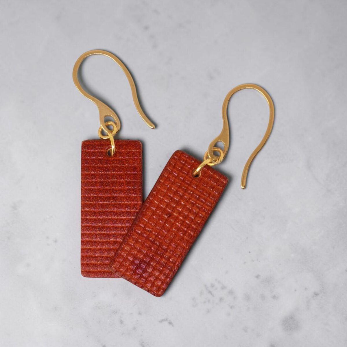 Boucles d'oreilles rectangulaires en cuir teinté cognac, texturage quadrillé, vues de face se chevauchant, suspendues à des boucles dorées sur fond gris