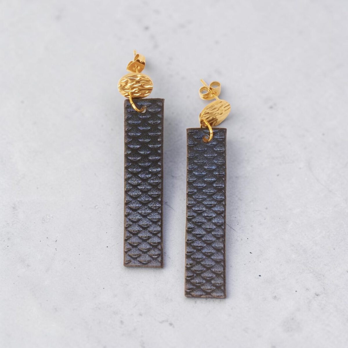 Boucles d'oreilles rectangulaires en cuir teinté noir charbon, texturage poisson, vues de face, suspendues à des boucles dorées rondes sur béton