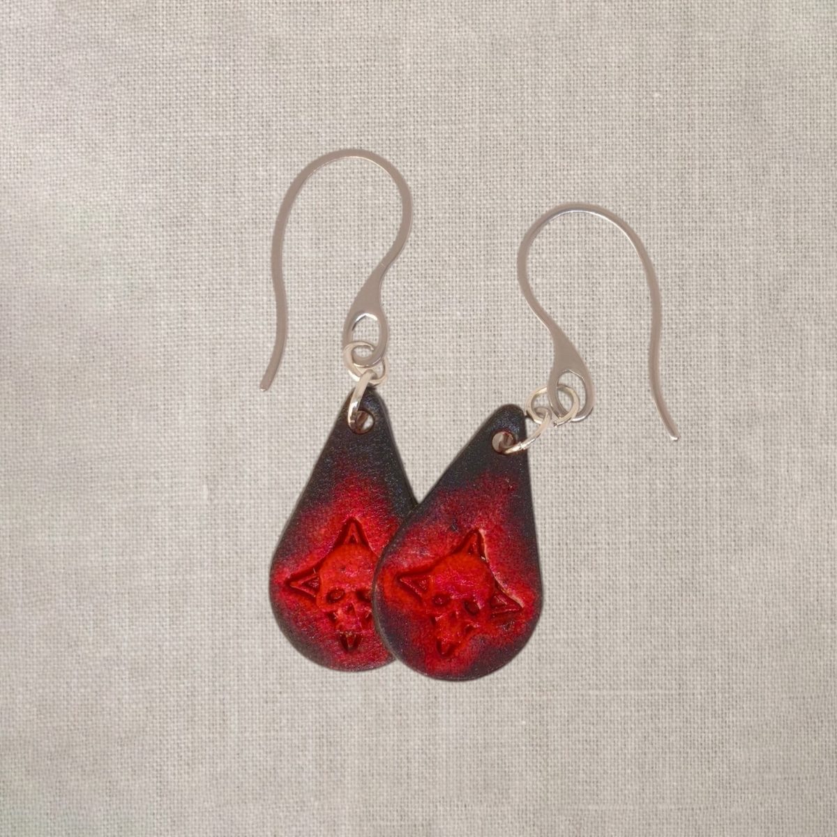 Boucles d'oreilles forme goutte d'eau en cuir teinture noire, estampages tête de mort teintés en rouge, vues de face se chevauchant, suspendues à des boucles argentées sur lin