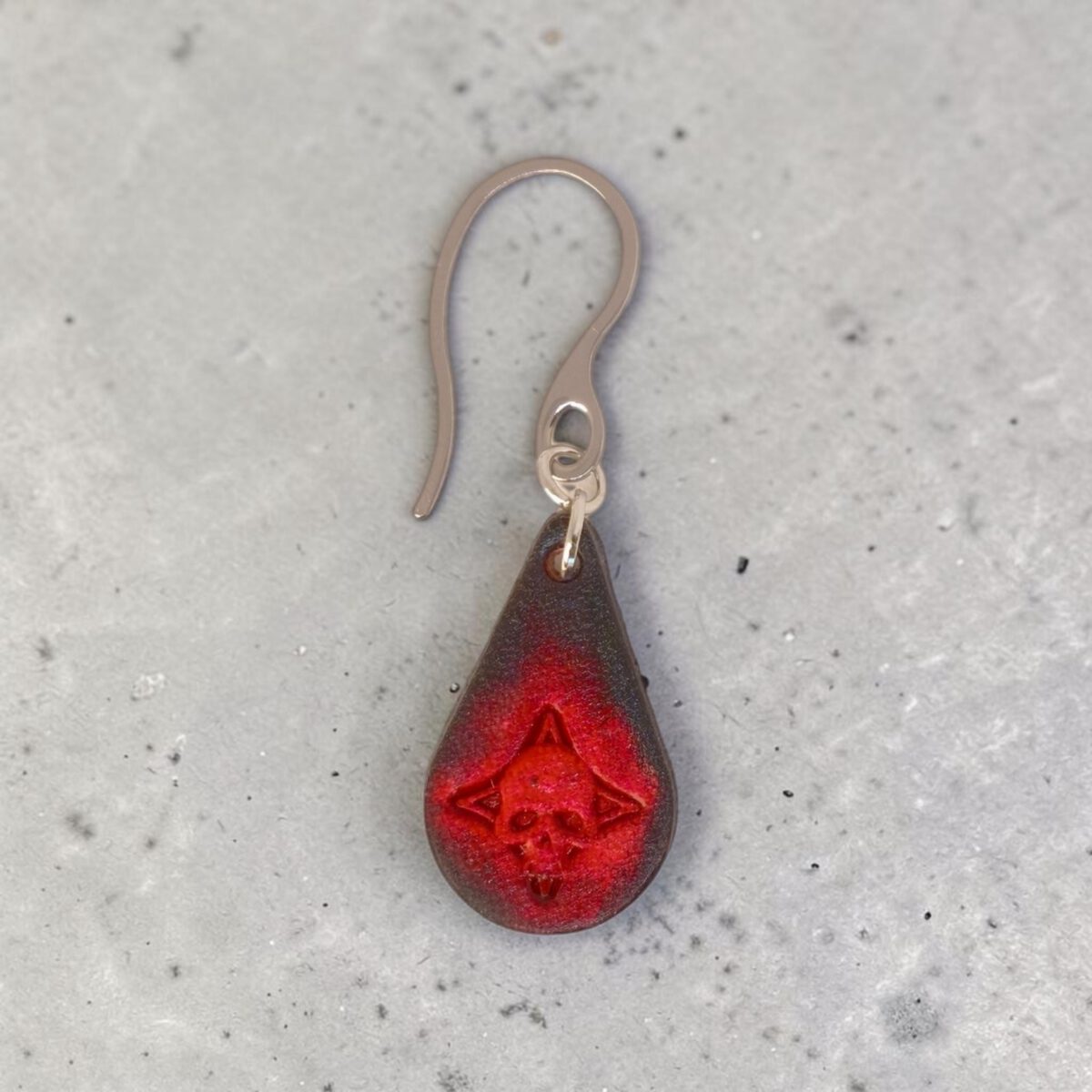 Boucle d'oreille forme goutte d'eau en cuir teinture noire, estampage tête de mort teinté en rouge, vue de face, suspendue à une boucle argentée, fond béton