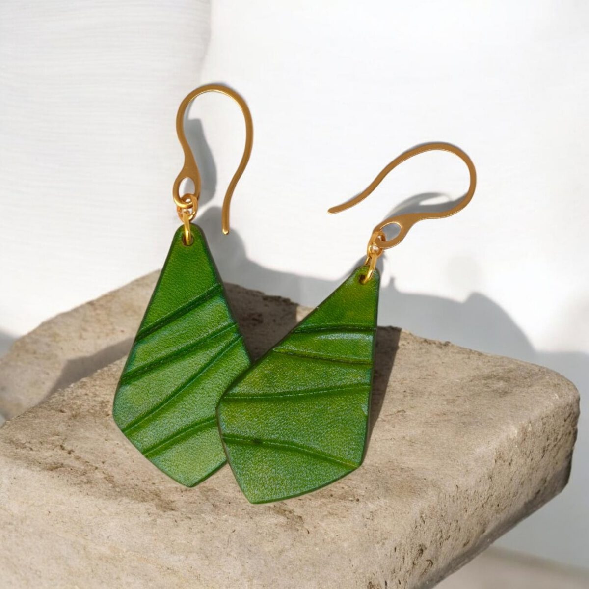 Boucles d'oreilles forme larme en cuir teinture vert forêt, finition galet, vues de face se chevauchant, suspendues à des boucles dorées sur pierre
