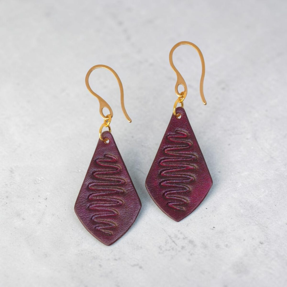 Boucles d'oreilles forme larme en cuir teinture violette, estampage "Lombard Street", vues de face, suspendues à des boucles dorées fond design béton