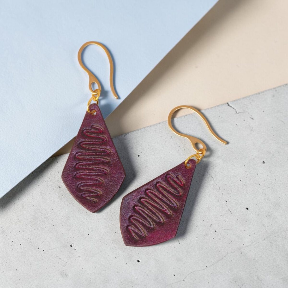 Boucles d'oreilles forme larme en cuir teinture violette, estampage "Lombard Street", vues de face, suspendues à des boucles dorées sur béton