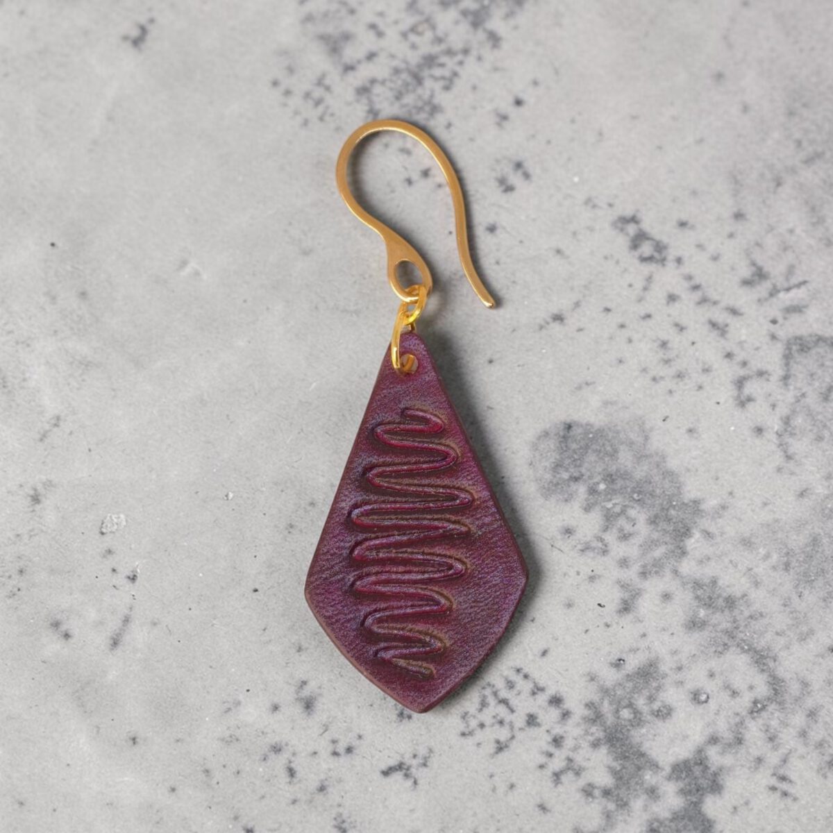 Boucle d'oreille forme larme en cuir teinture violette, estampage "Lombard Street", vue de face, suspendue à une boucle dorée, sur béton