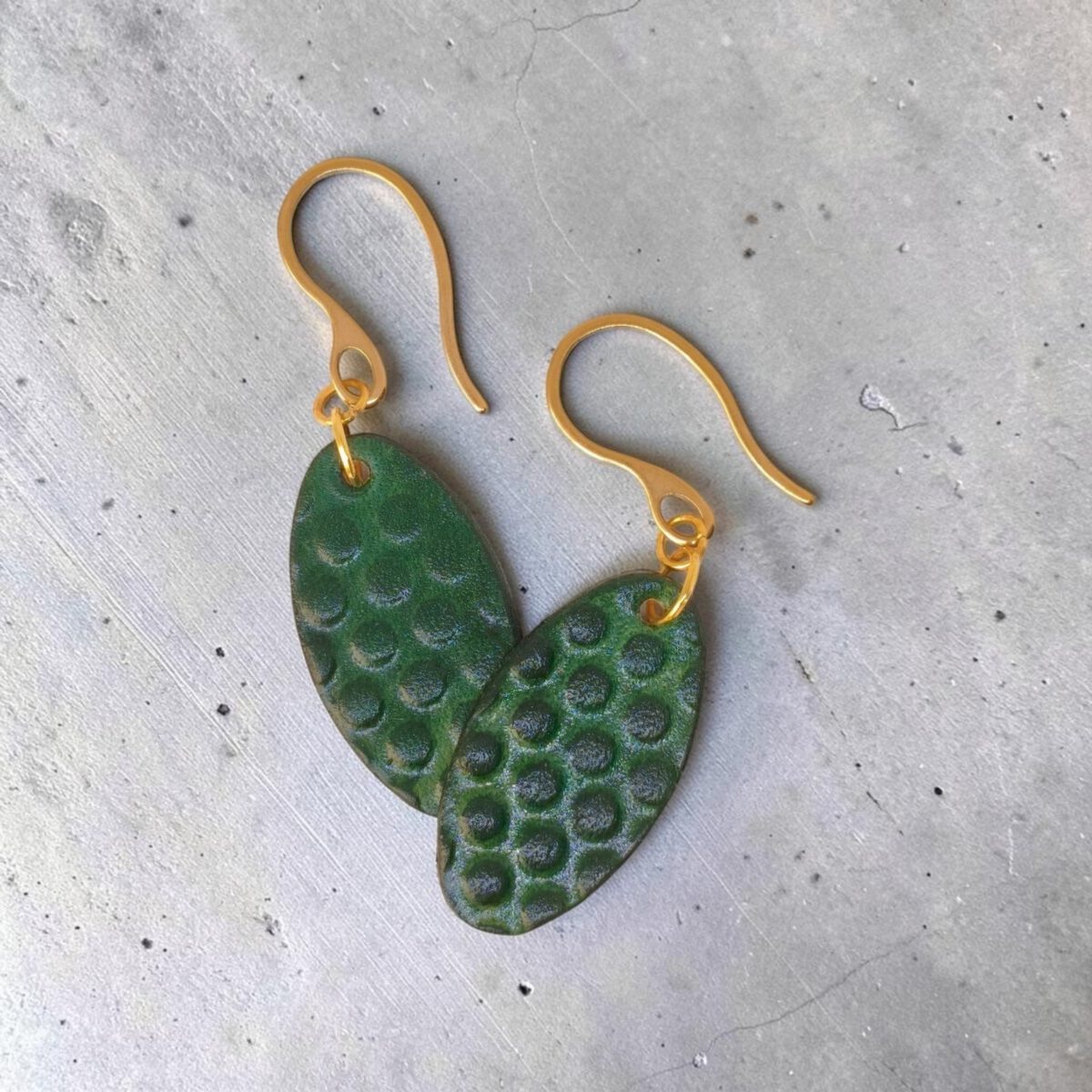 Boucles d'oreilles ovales en cuir teinte vert forêt, texturage ronds, vues de face se chevauchant, suspendues à des boucles dorées sur béton